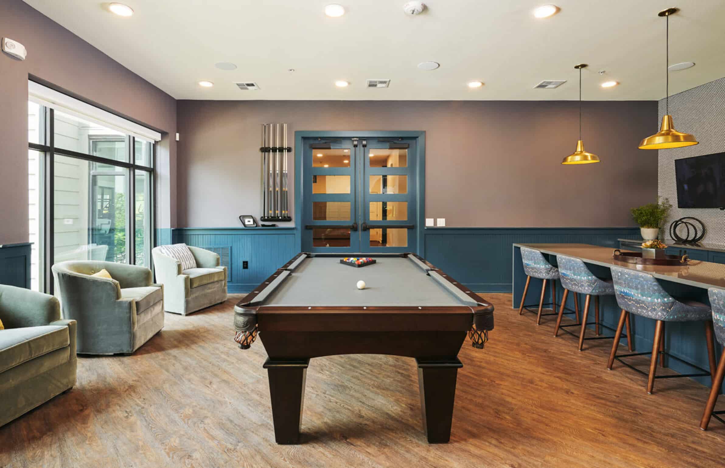 Modern game room at The Copeland with a pool table, bar seating, armchairs, and large windows letting in natural light.