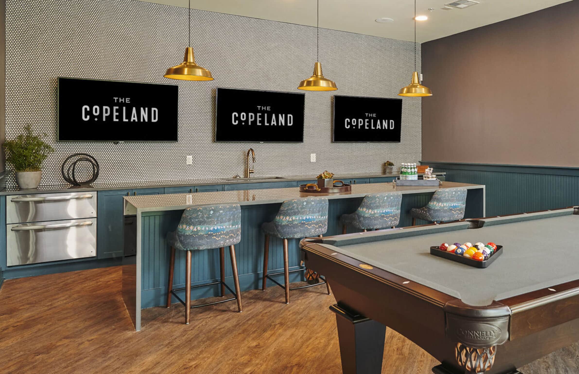 Modern game room at The Copeland with a pool table, bar seating, three TVs, and gold pendant lights above the counter.