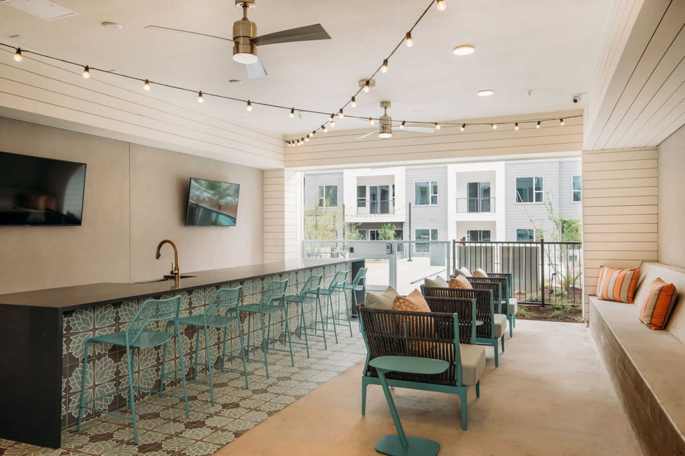 Modern outdoor lounge at The Copeland with a bar, teal chairs, string lights, TV, and cushioned seating overlooking apartments.