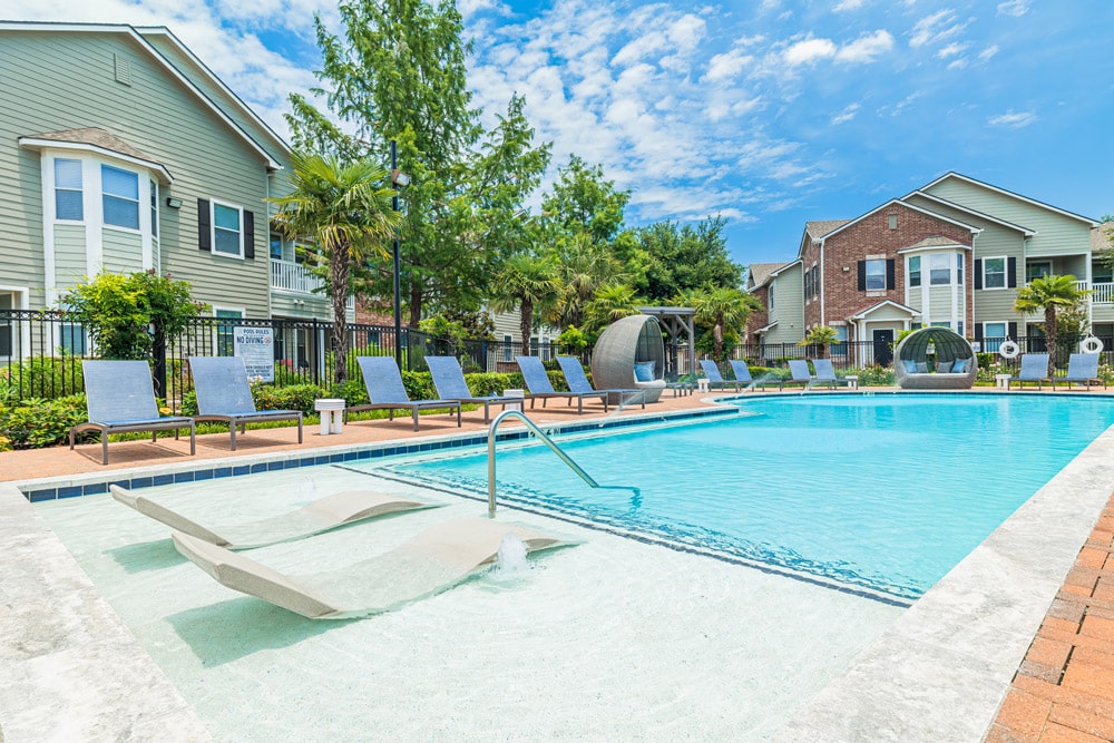Outdoor swimming pool at The Bryx at Kingwood with lounge chairs, sunbeds in shallow water, and buildings in the background.