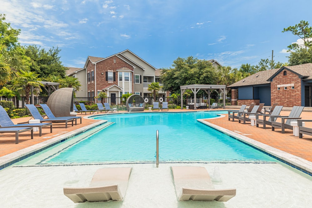 The pool at The Bryx at Kingwood with lounge chairs, sunbeds, and brick buildings in the background on a sunny day.