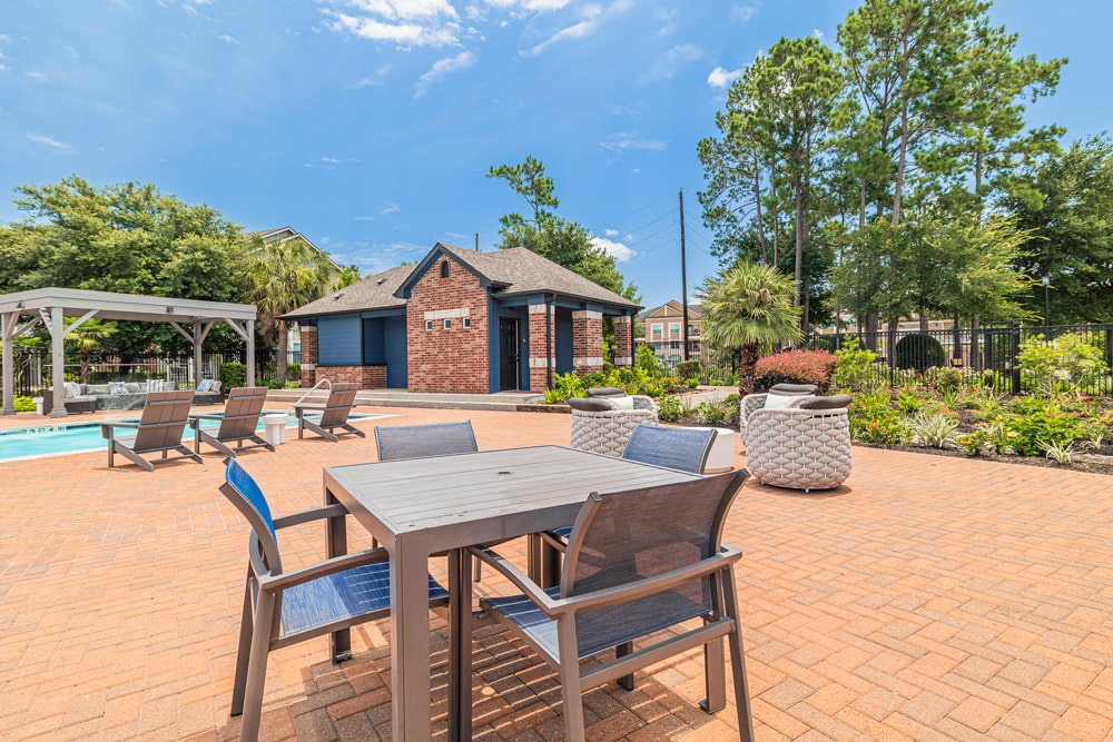 Outdoor patio at The Bryx at Kingwood with table, chairs, lounge chairs, pool, brick building, and trees under a sunny sky.