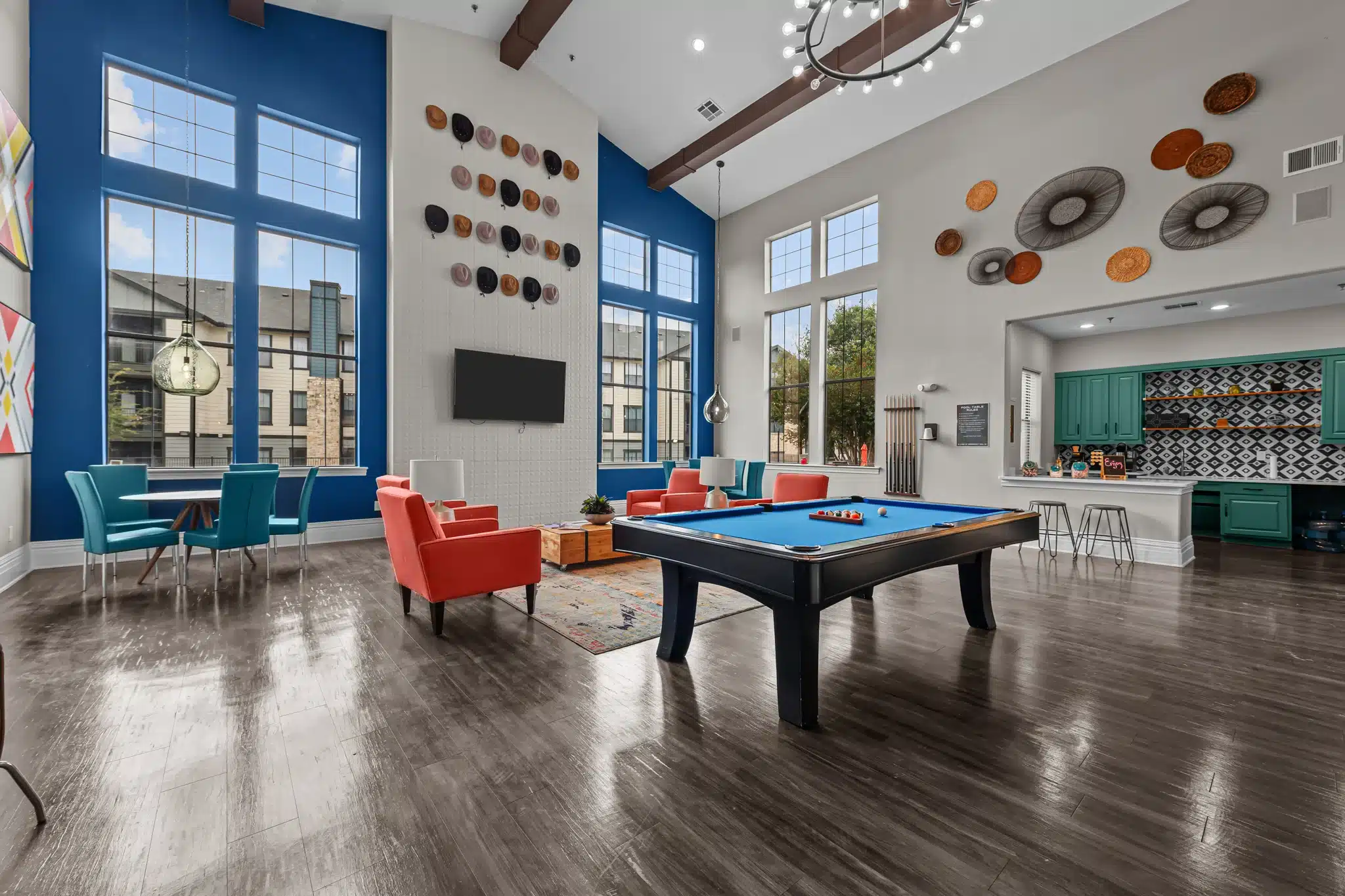 Modern game room at The Heritage with pool table, colorful seating, wall art, large windows, and a kitchenette in the back.