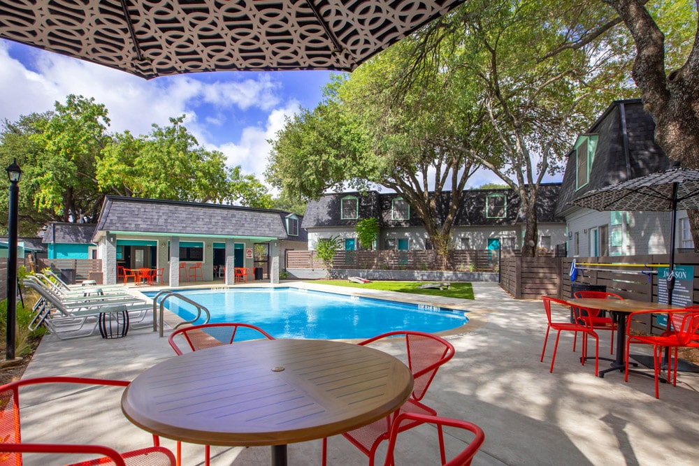 The Jackson pool area with red chairs and tables, lounge chairs, and trees providing shade on a sunny day.