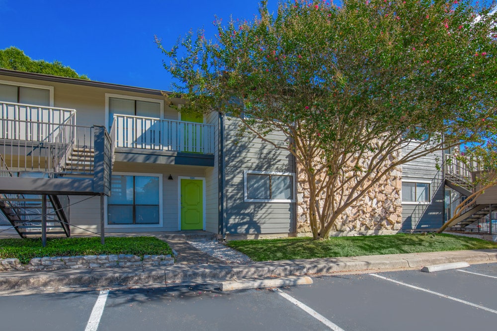 Two-story apartment building at The Escapade with green front doors, a balcony, and large trees in front.