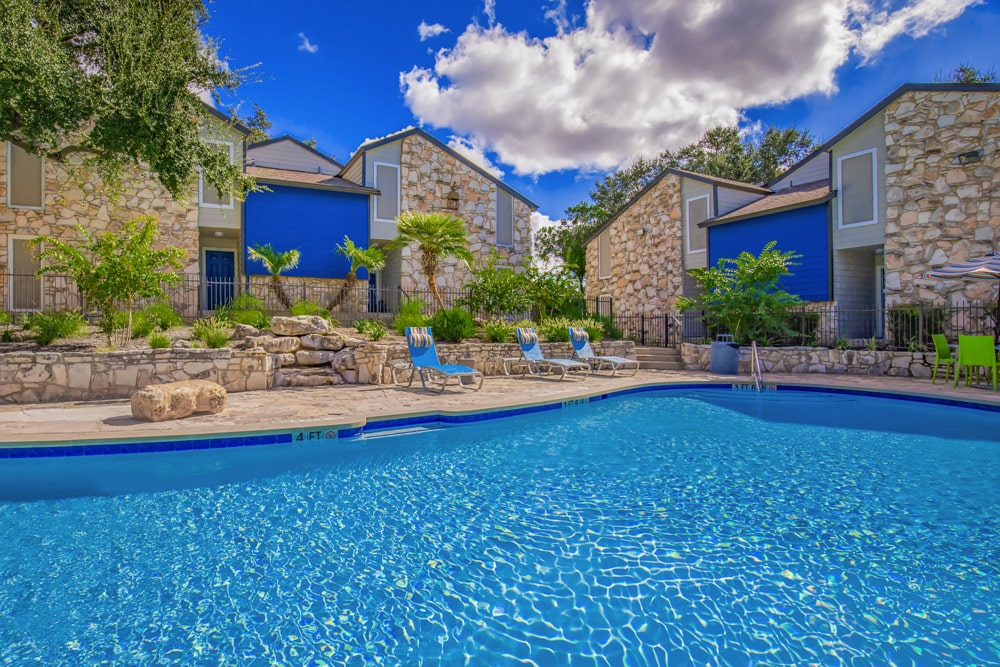Outdoor swimming pool  at The Escapade with lounge chairs, and stone buildings with blue accents under a bright sky.