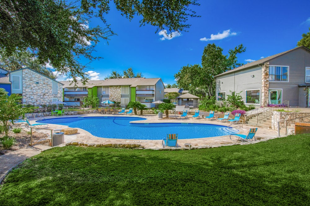 Sunny outdoor pool area at The Escapade with lounge chairs, surrounded by grass, trees, and apartment buildings.