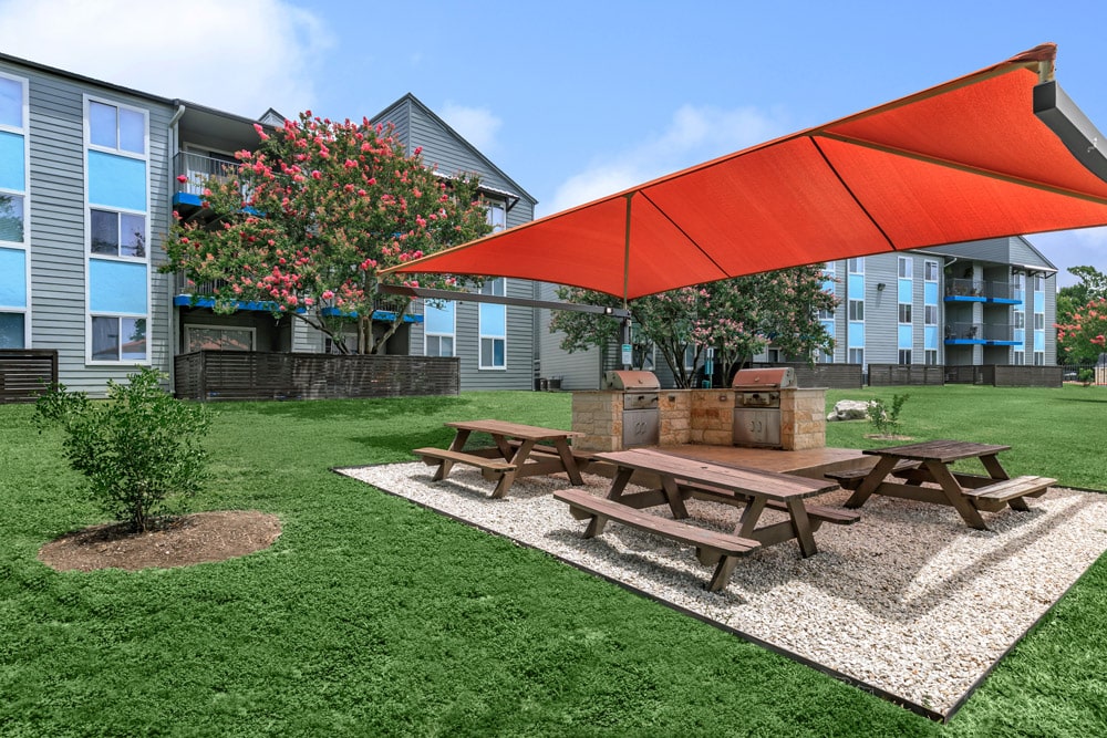 Outdoor picnic area at The Keller with wooden tables, grills, and red shade canopy on a sunny day.