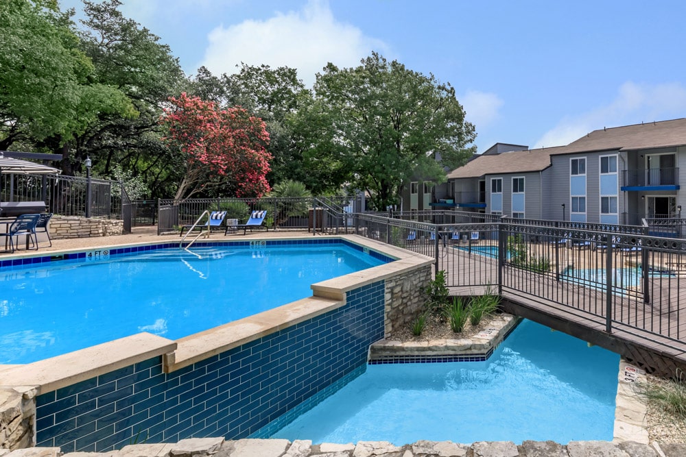 Outdoor swimming pool at The Keller with a stone deck, lounge chairs, and nearby apartment buildings surrounded by trees.